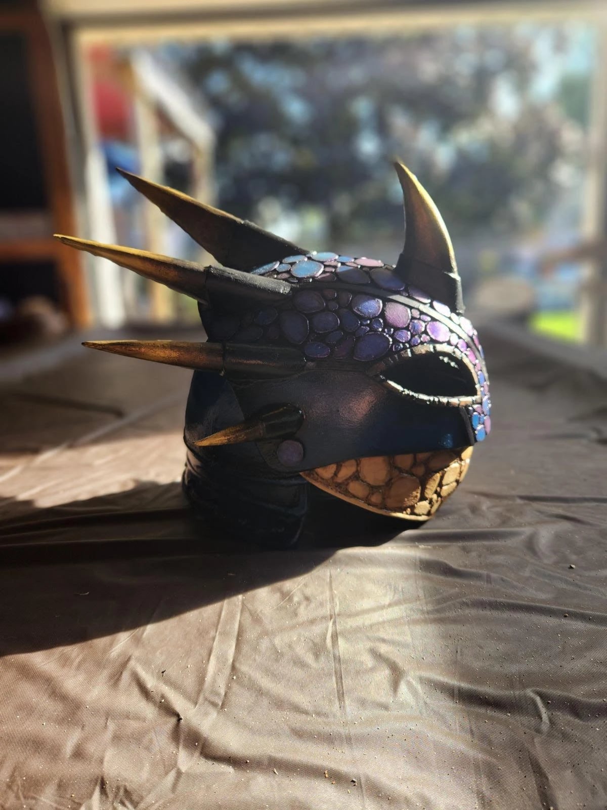 Horned helmet study on a table in sunlight: dark scaled surface painted with iridescent blues and purples, gold spikes, and a gold-scaled jaw guard.