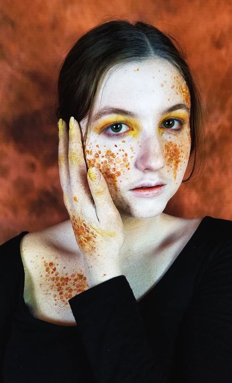 SFX makeup portrait: a performer with amber honeycomb-textured prosthetic transfers across one cheek, hand, and chest, against a rust-coloured backdrop.